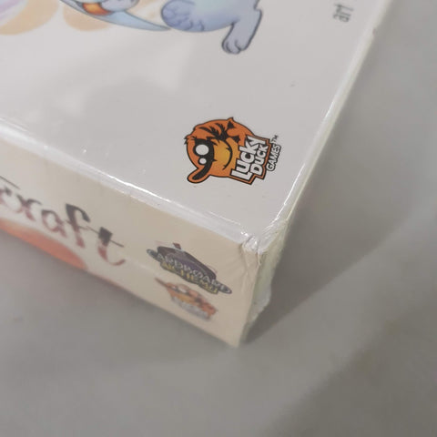 A close-up of a Flamecraft (Damaged) board game box corner, wrapped in plastic. The Lucky Duck Games logo—an orange duck with sunglasses—is visible, and there is some light wear on the corner.