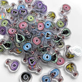 A vibrant pile of Lux Lupo’s Apothebakery: The Master Alchemist Expansion acrylic pins—potion bottle shapes with whimsical eyes, paw prints, and symbols—lies scattered like upgraded tokens on a white surface.