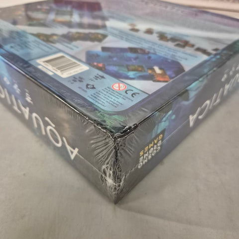 A shrink-wrapped Cosmodrome Games Aquatica EN/DE (Damaged) board game box, themed around underwater kingdoms, with a crushed corner, lies on a light-colored surface.