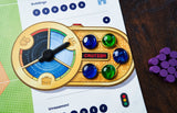 A close-up of a game component from Capstone Games’ Bus: Complete Edition shows a gold circular dial with a black pointer, four glass marbles in various colors, purple tokens, and wooden pieces on the public transport-themed board.