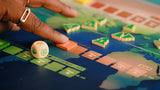A close-up of a hand moving a piece on Daybreak, a colorful climate action board game by CMYK featuring world maps, green tree tokens, a symbolic white die, and multicolored tiles.