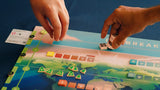 Two hands move pieces on Daybreak, a colorful world map board game by CMYK. Tokens, cubes, and cards are arranged as players cooperate to achieve net-zero emissions through climate action gameplay.