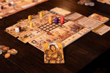 A close-up of Garphill Games' "Ezra and Nehemiah" board game in play on a dark wood table, showing colorful wooden pieces, illustrated cards—one with Nehemiah rebuilding the temple—and numbered tokens.