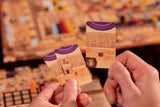 Two hands hold up Ezra and Nehemiah cards by Garphill Games, featuring brick wall and purple curtain designs with a box and lock, set against a blurred board game scene with pieces in the background.