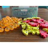 Orange marbles and colorful wooden fish are scattered on a table in front of a Finspan Bundle box by Stonemaier Games, capturing the excitement of ocean exploration in the Finspan board game.