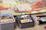 Close-up of stacked square tiles from Great Western Trail: El Paso by Lookout Games, with the top tile featuring cattle trade icons and artwork. A blurred game board appears in the background.