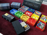 The Hegemony - 3D Upgraded Components by Game Maker Team are shown in organized, color-coded trays with gaming tokens, cards, and pieces on a red surface, with the board game box in the background.