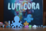 A blue wooden octopus game piece stands before other colorful animals, capturing the lively underwater theme of Lumicora by Pegasus Spiele, with the game's blurred box in the background.