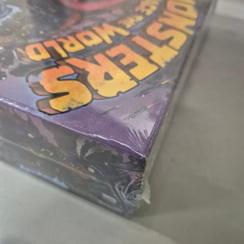 A close-up shows slight damage to the cardboard corner of a shrink-wrapped Monsters Menace the World (Damaged) board game by Renegade Game Studios, with bold yellow and orange title lettering visible on the box.
