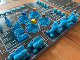 A close-up of Rise of the Wastelands Deluxe Kickstarter Edition by Wulfhorn Games shows blue miniatures, buildings, and vehicles on a post-apocalyptic player mat, with colored cubes tracking resources among ruined cities.
