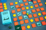 The Oink Games board game Souvenirs from Venice is set up on a gray surface with orange and blue square tiles for collection, yellow round tokens, a green die, and a light blue box.