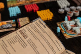 A close-up of The Anarchy rulebook by Renegade Game Studios, open on a dark table with colorful game tokens and pieces scattered around.