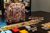 A tabletop setup for The Anarchy by Renegade Game Studios, showing the game box with medieval artwork and an array of colorful pieces, cards, and tiles neatly arranged on a dark table.