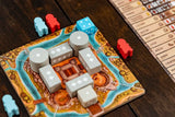 A close-up of The Anarchy by Renegade Game Studios, a medieval strategy board game with a castle, red and blue wooden meeples, a blue die with white stars, and a player reference card on a wooden table.