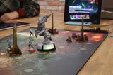 A tabletop game scene features Pegasus Spiele’s Teburu Game Platform Bundle with miniatures and 3D terrain on a vibrant board, while a tablet shows Teburu details in the background and a person’s arm appears on the left.