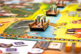 A close-up of Tianxia by Board & Dice, a colorful strategy board game set in the Warring States period, with a wooden boat on a river surrounded by cards, tiles, and tokens atop illustrated terrain with paths and water.