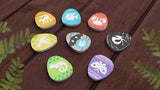 Eight colorful, egg-shaped tokens with animal silhouettes from Bad Comet's Wondrous Creatures sit on a wooden surface among green fern leaves—ideal for strategic board games with worker placement and tableau-building mechanics.