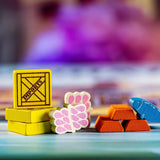 Yokohama: Premium Tokens by Synapses Games features colorful wooden accessories: yellow IMPORTS crates, pink grapes, orange gold bars, and a blue fish—ideal upgrades for your game, set against a vibrant blurred background.